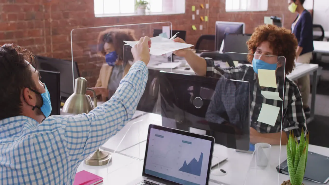 Diverse business colleagues exchanging documents over sneeze screen wearing face masks