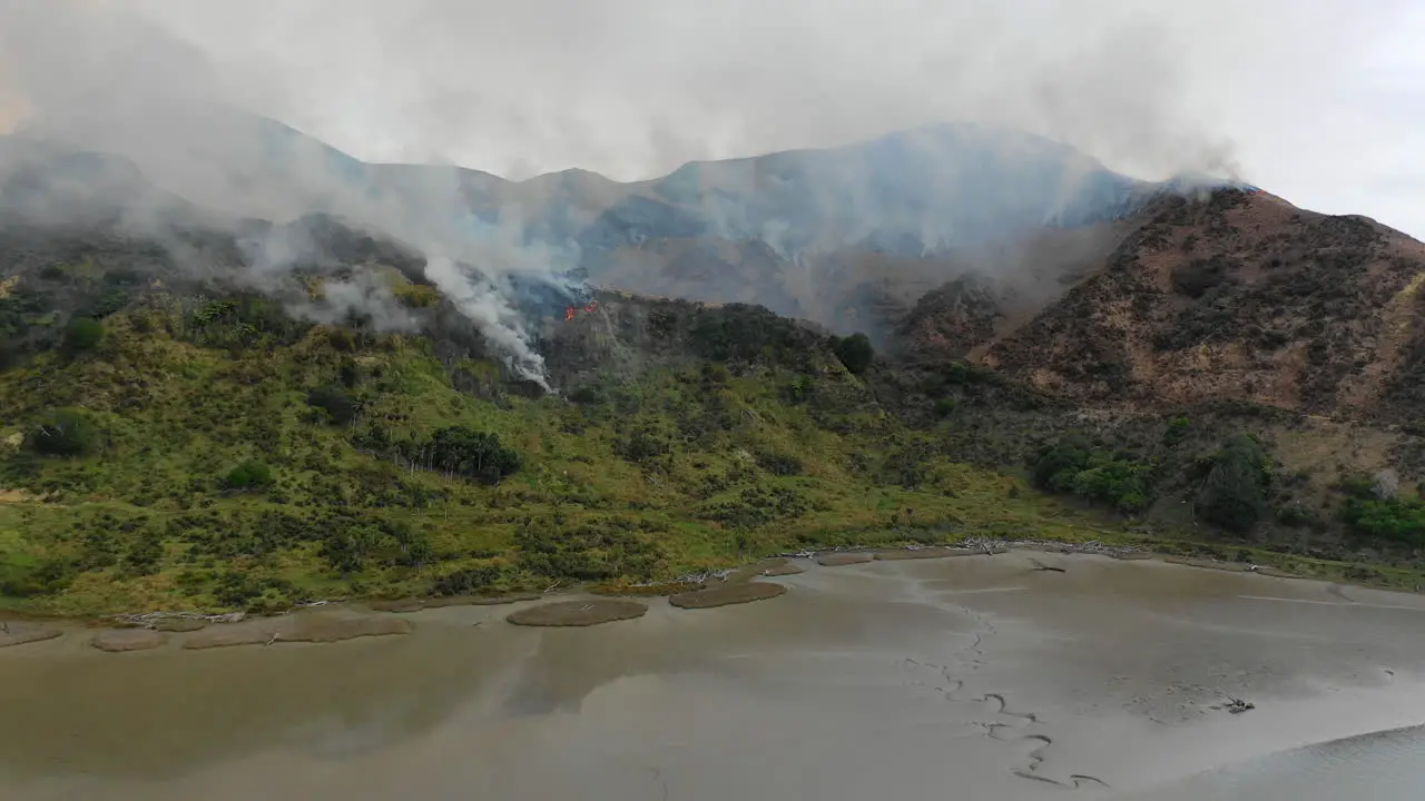 Aerial view flying over a mud flat toward a wildfire burning the mountainside on New Zealand's North Island