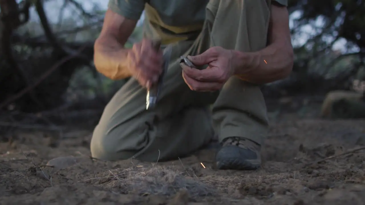 Caucasian male survivalist using knife and flint to spark fire with tinder at camp in wilderness