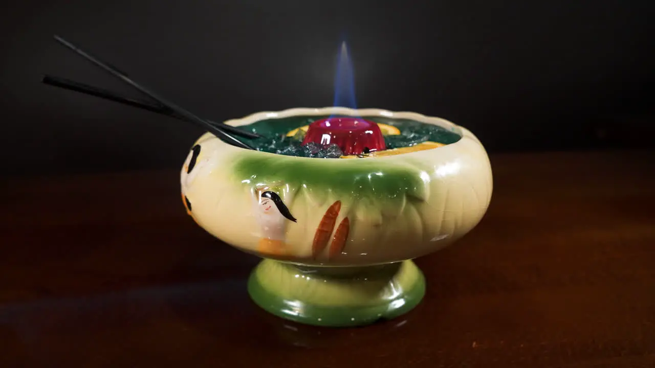 Blue flame dances in the center of a shareable tropical tiki style cocktail with floating orange slices slider orbit 4K