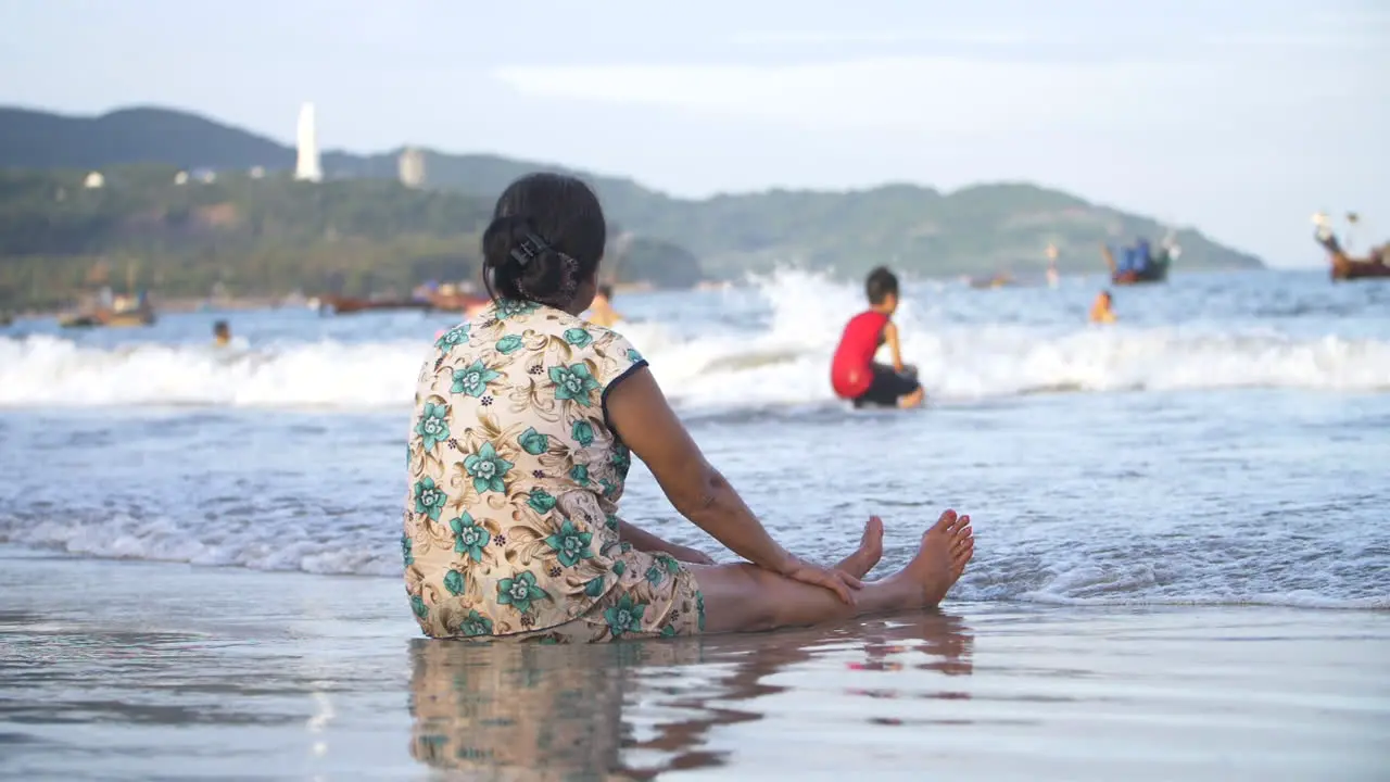 Woman Sat at Beach Shore Line watching her Children Play in Ocean