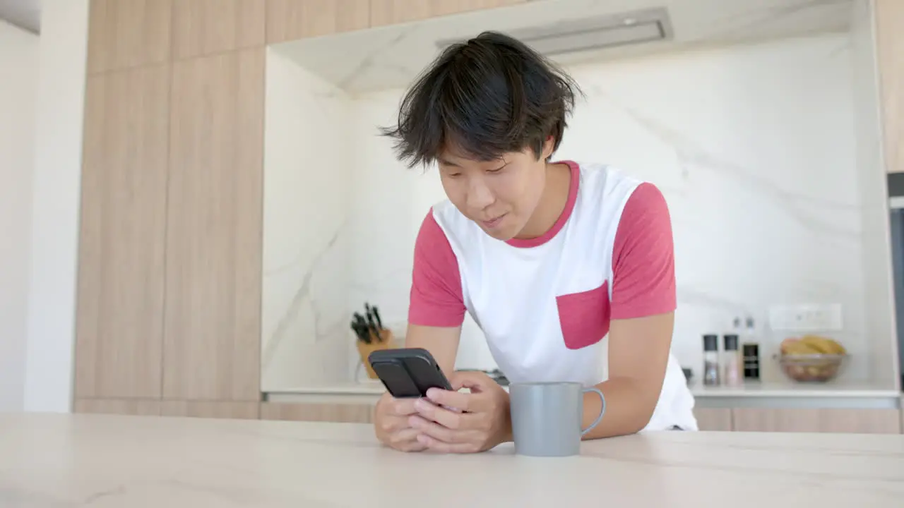 Teenage Asian boy smiles while using his smartphone at home in the kitchen