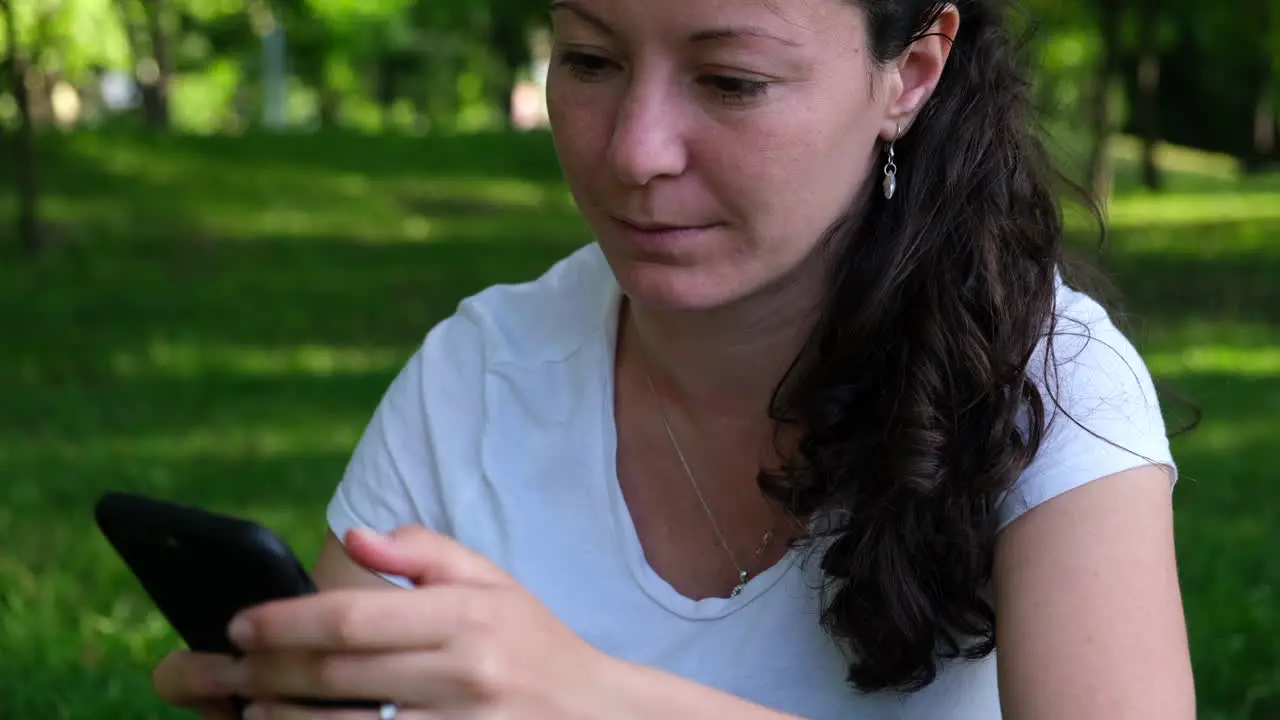 Medium shot of a young woman sitting on the grass in the park scrolling through social media on her smartphone and laughing