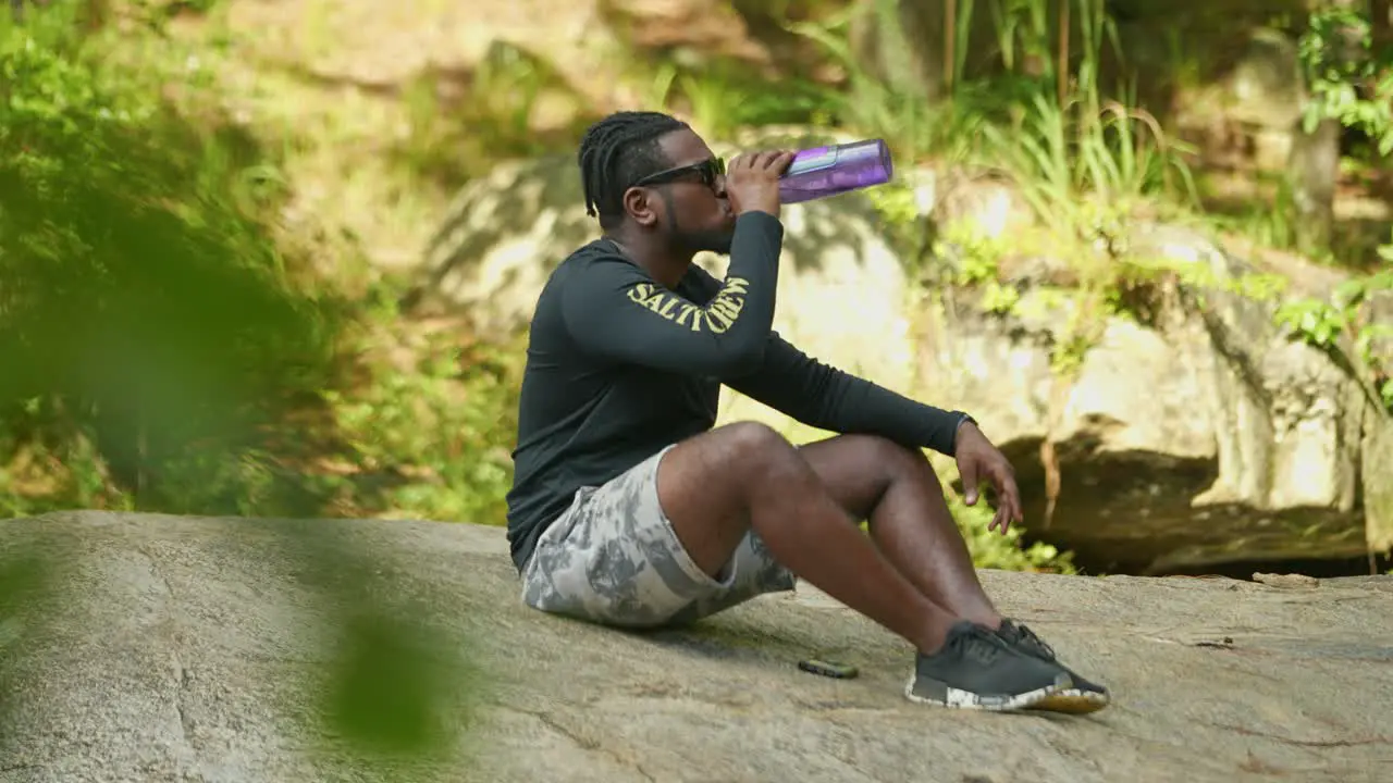 A man takes a sip of water on a giant rock in the middle of the woods on a bright sunny day