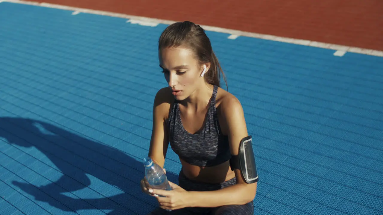 Tired Fitness Woman Sitting At Sport Court With Bottle Of Cold Water Wiping Sweat From Forehead And Resting After Workout