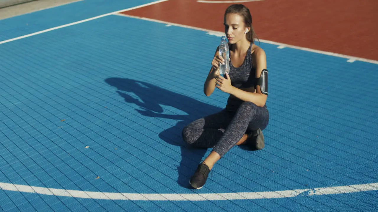 Fitness Woman Sitting At Sport Court And Drinking Cold Water After Workout On A Summer Day