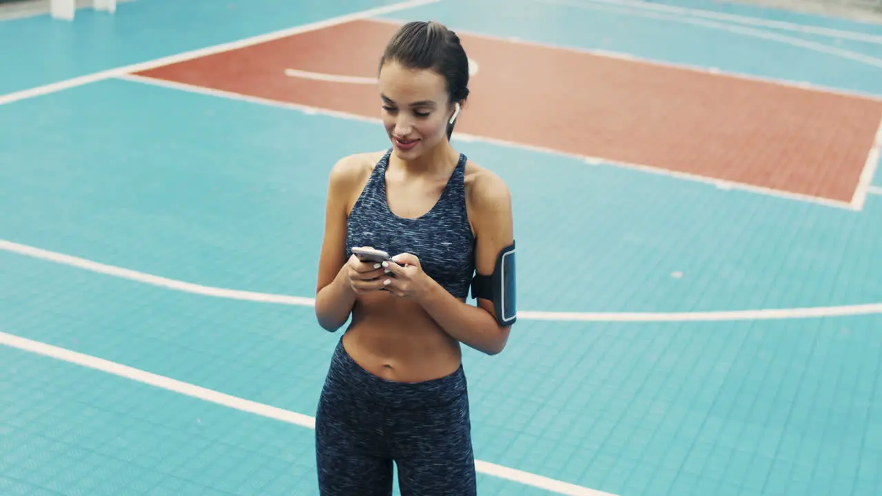 Sporty Girl With Airpods Texting Message On Smartphone While Standing At Outdoor Court On A Summer Day