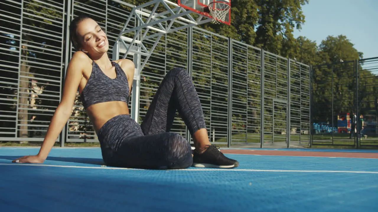 Happy Fit Girl With Airpods Sitting At Sport Court In Summer Sunlight And Resting After Workout