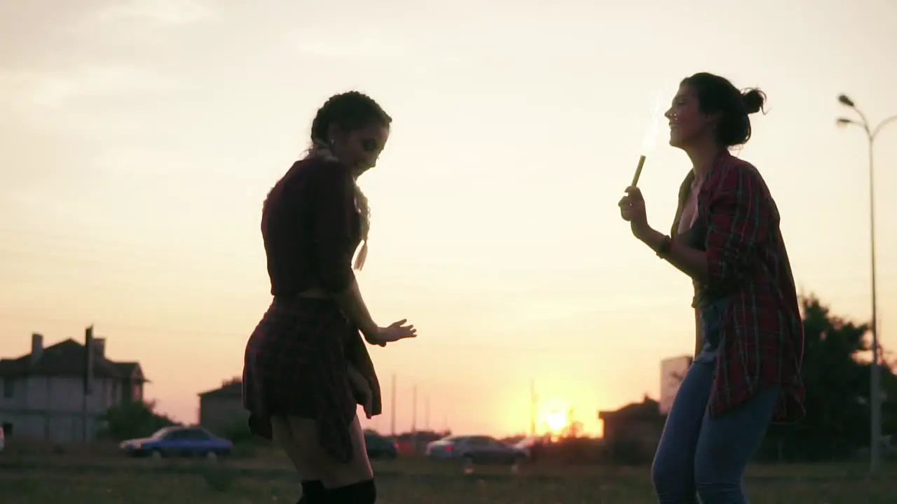 Two Attractive Girlfriends Dancing Together And Waving With Firework Candles During Sunset