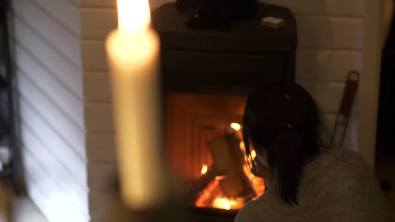 Woman stares thoughtfully into flames of home fireplace candle flickers in foreground