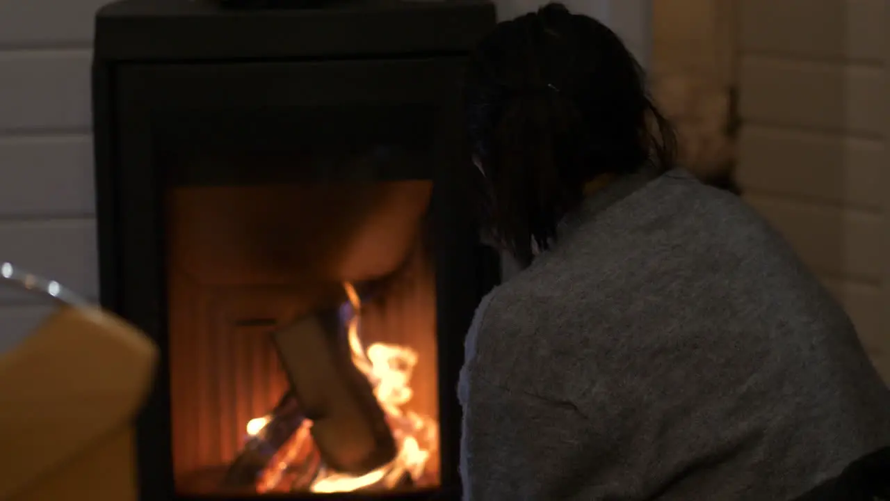 Woman thoughtfully watches flames burning logs in indoor fireplace