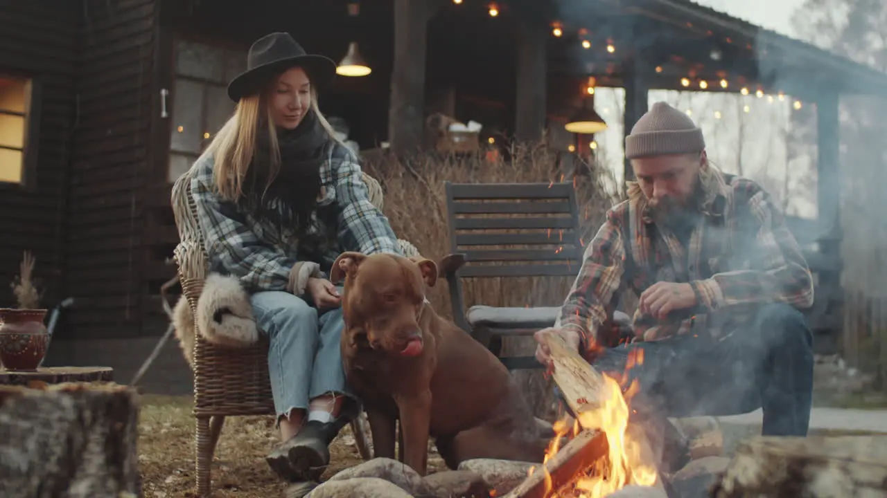 Young Couple and Dog Sitting by Bonfire near Farmhouse