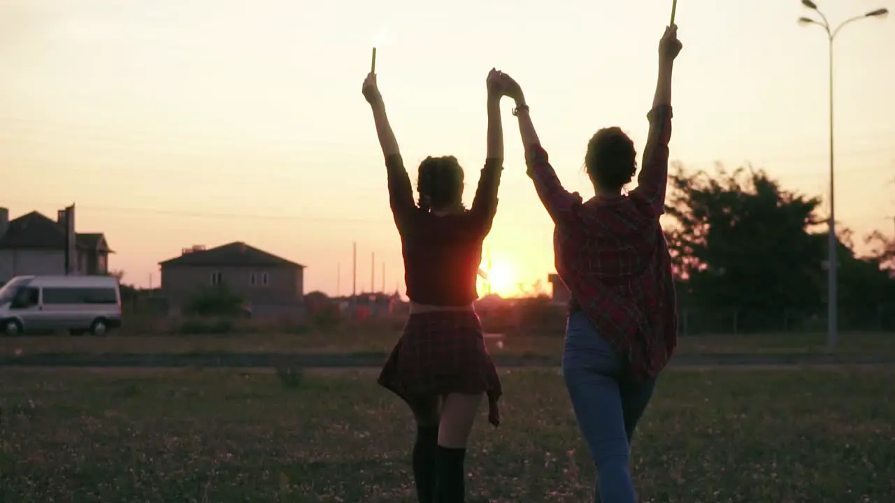 Back View Of Two Girlfriends Walking Forward And Waving With Firework Candles During Sunset