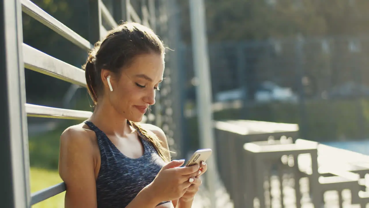 Sporty Girl With Airpods Laughing While Texting Message On Smartphone After Workout At Outdoor Court On A Summer Day