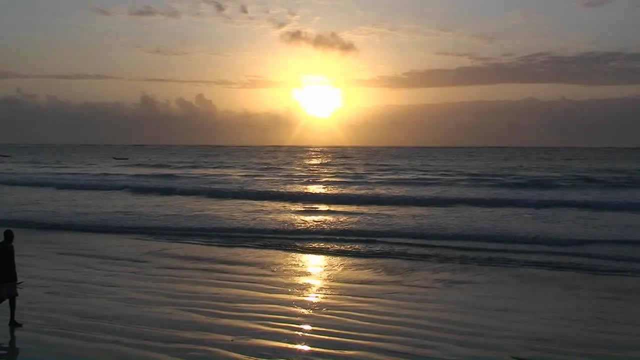 A man walks along the beach carrying a stick as the waves come in and the sun sets