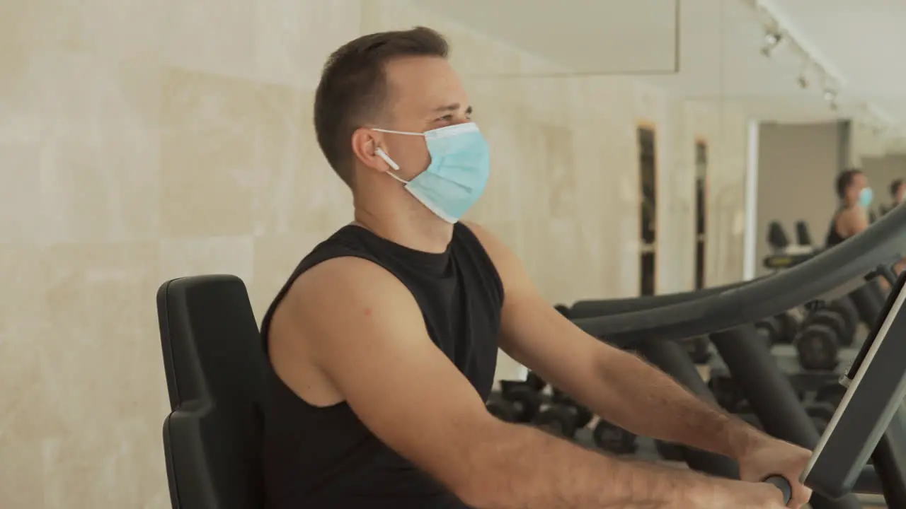 Young Strong Man With Face Mask Uses A Smartphone And An Exercise Machine In The Gym