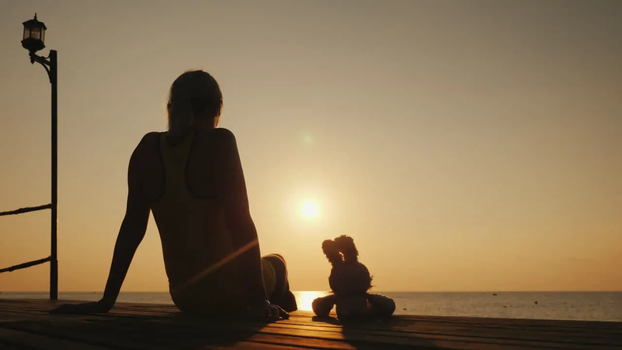 A Woman Sits On A Pier Next To A Toy Hare Together They Meet The Sunrise Over The Sea Romantic Mood 