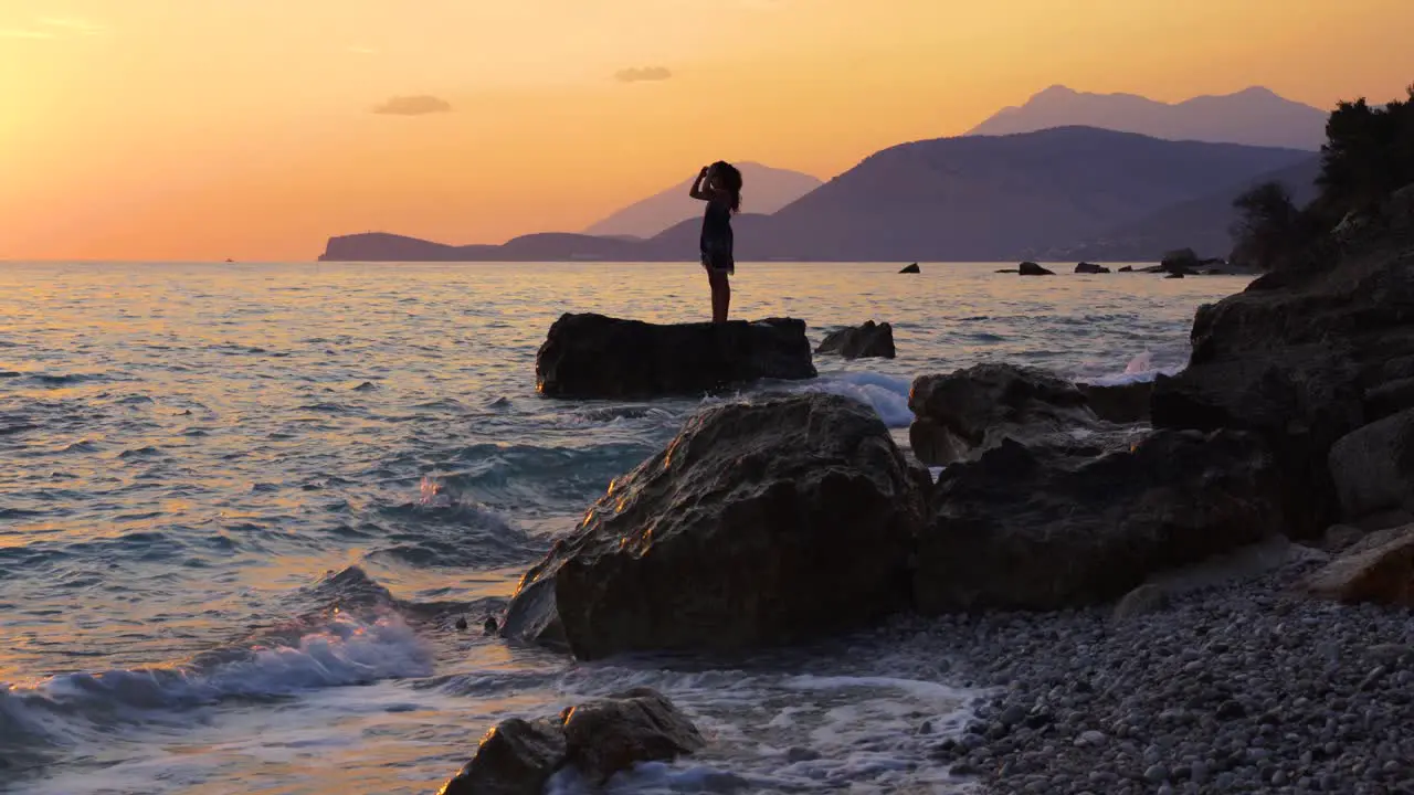 Girl standing on cliff washed by sea waves at beautiful sunset with silhouette of mountains and orange sky