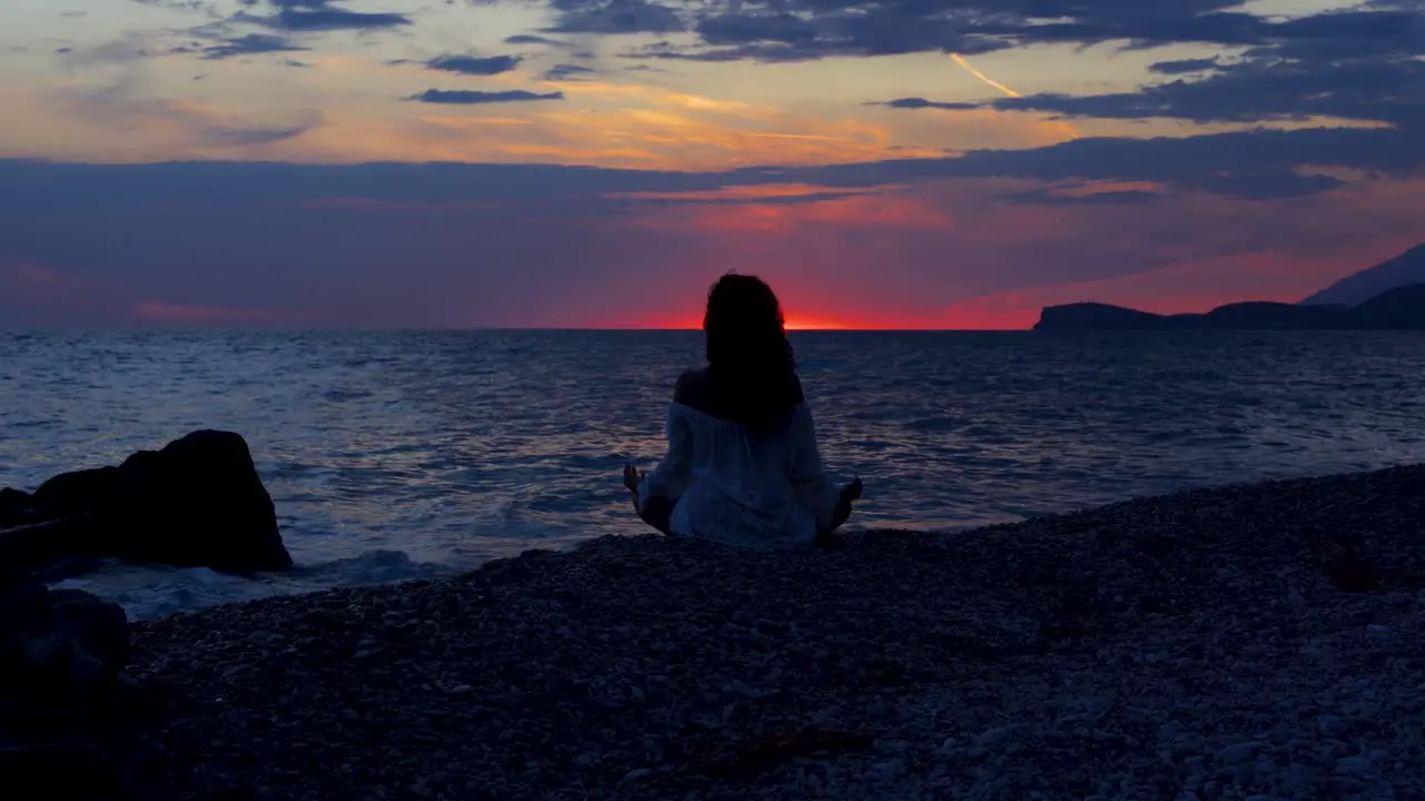 Yoga pose of girl sitting on pebbles of tranquil beach washed by sea reflecting colorful sunset