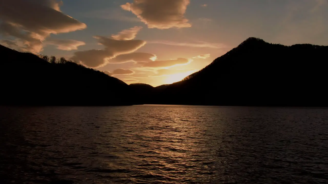 High contrasted aerial low shot flying over a mountain lake towards the sun and beautiful clouds peaceful serene