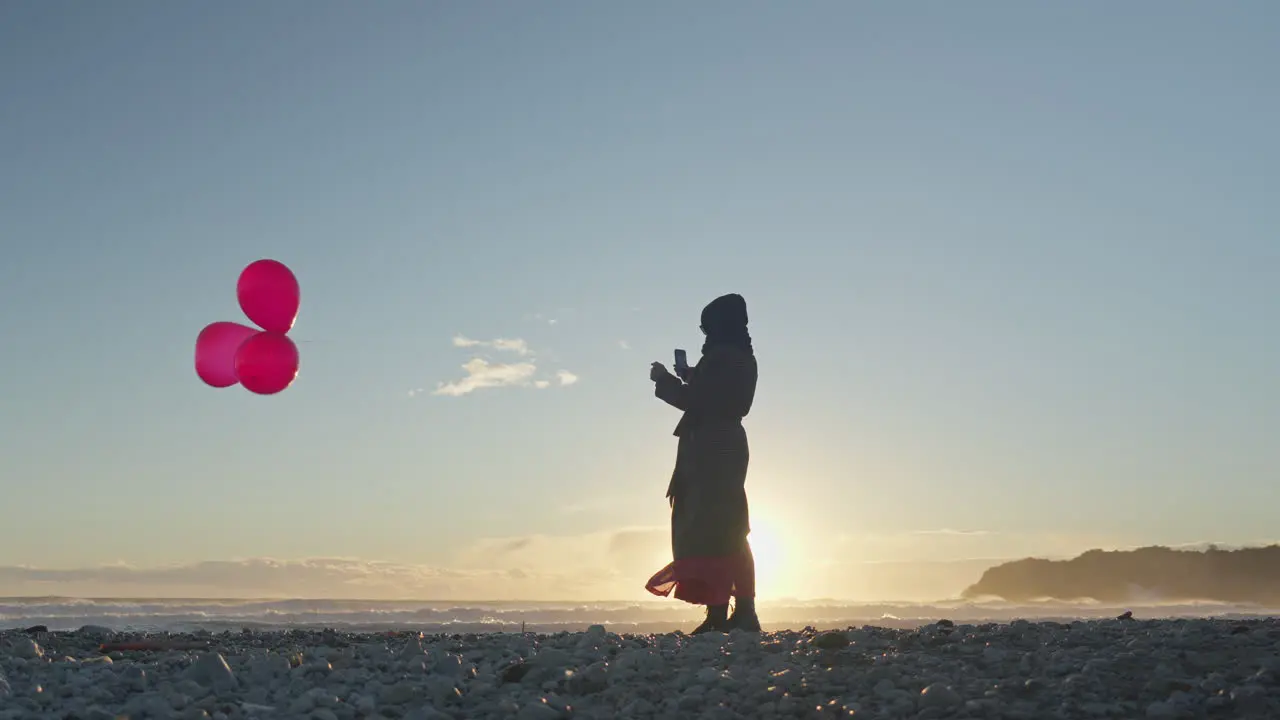 A woman's silhouette is holding three red balloons at the beach as the sun sets