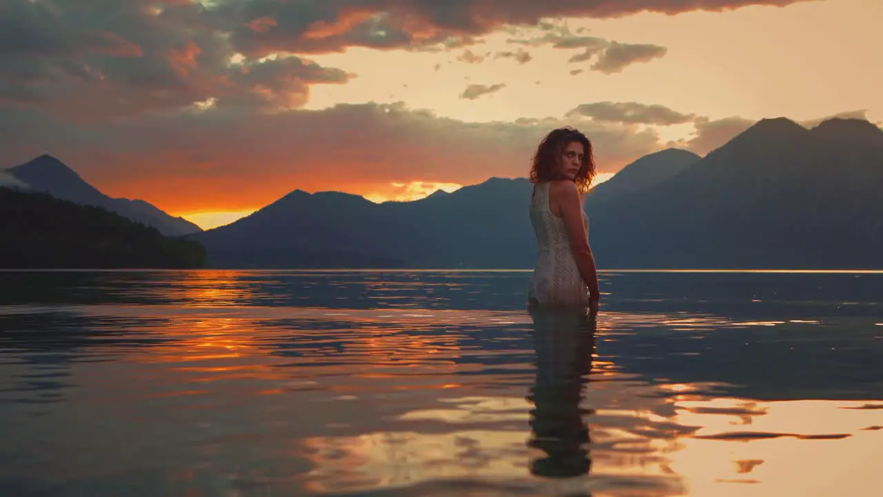 Atmospheric Cinemagraph Seamless video loop of a beautiful young model woman standing in the waves of lake Walchensee in Southern Germany-Bavaria in a white fashion dress by sunset facing the sun