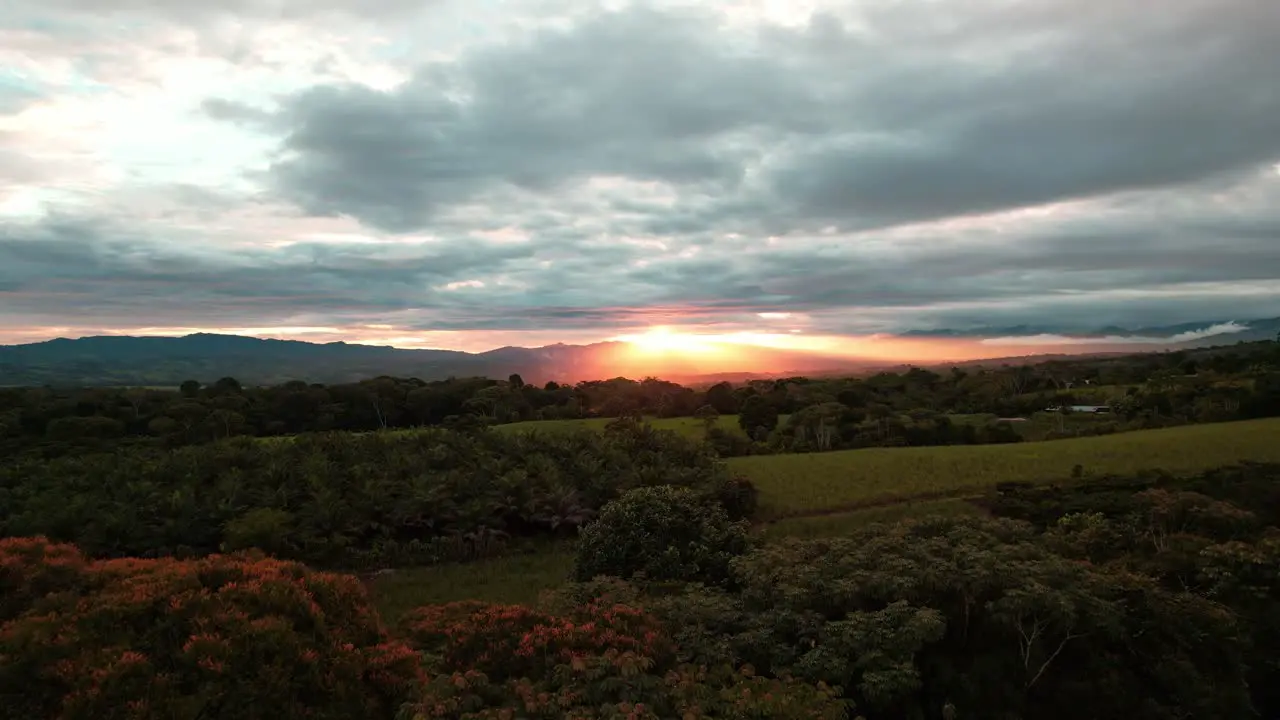 Aerial dolly in flying over dense woods and green valley with mountains in background on a cloudy sunset