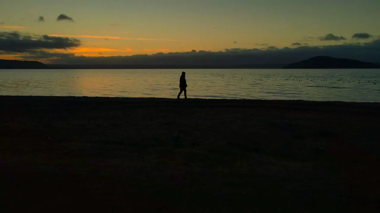 Slow pan to a Lonely Person at Dusk slowly walking alone along shore at sunset with horizon in background