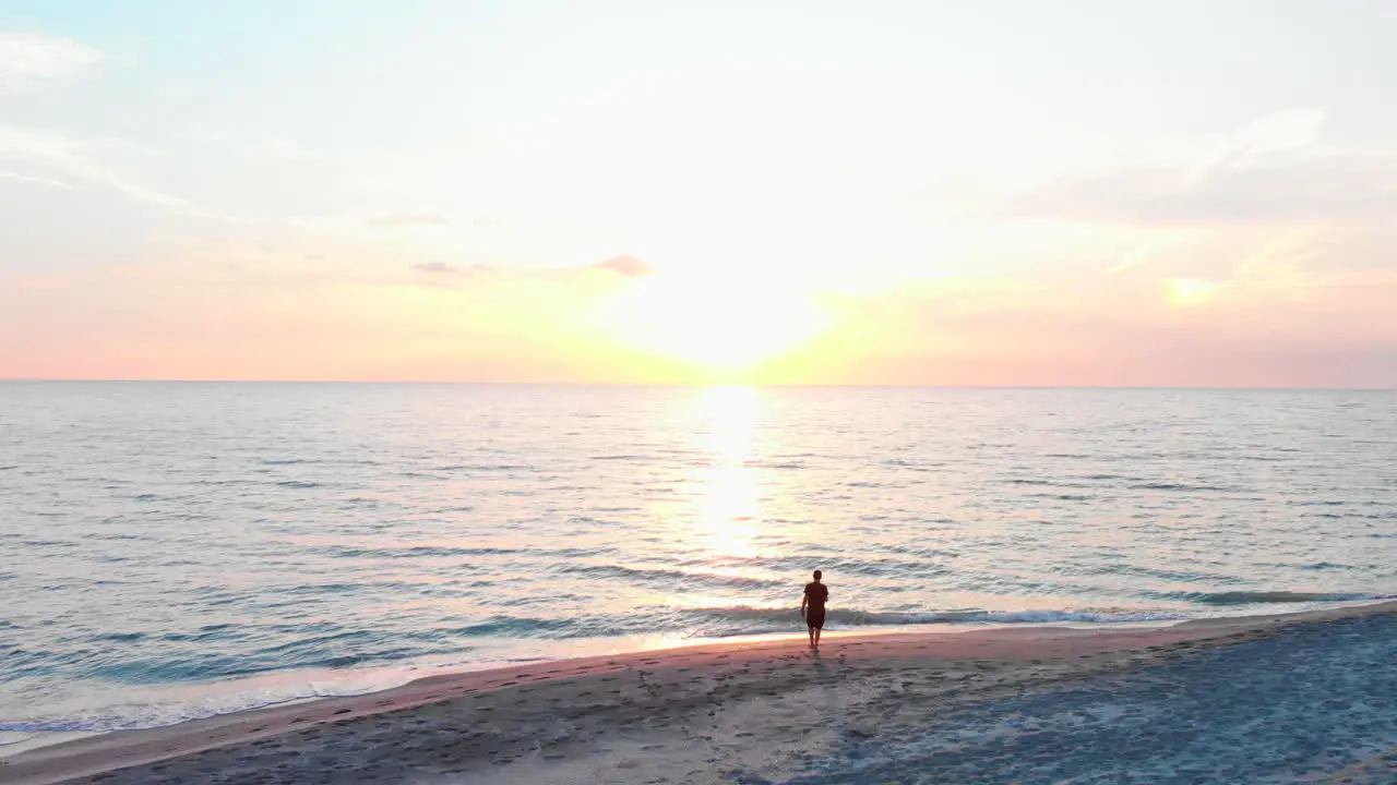 Aerial adult male standing on beach watches sunrise slow motion flyover