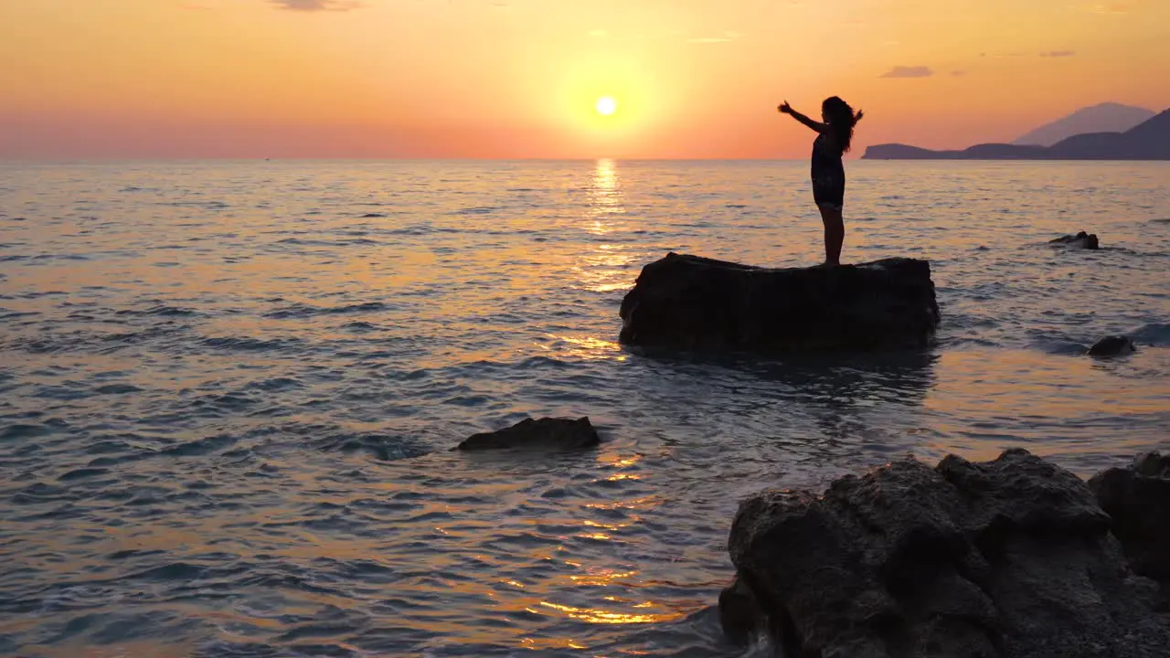 Yoga exercising performed by young woman standing on rock washed by sea with orange sky of sunset background