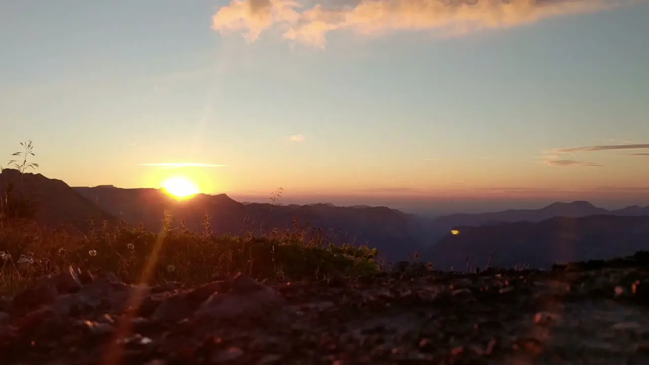 Timelapse sunset in alpine region orange sun is setting on horizon grass and flowers blowing in the wind camera is at low angel near the ground clouds on the sky