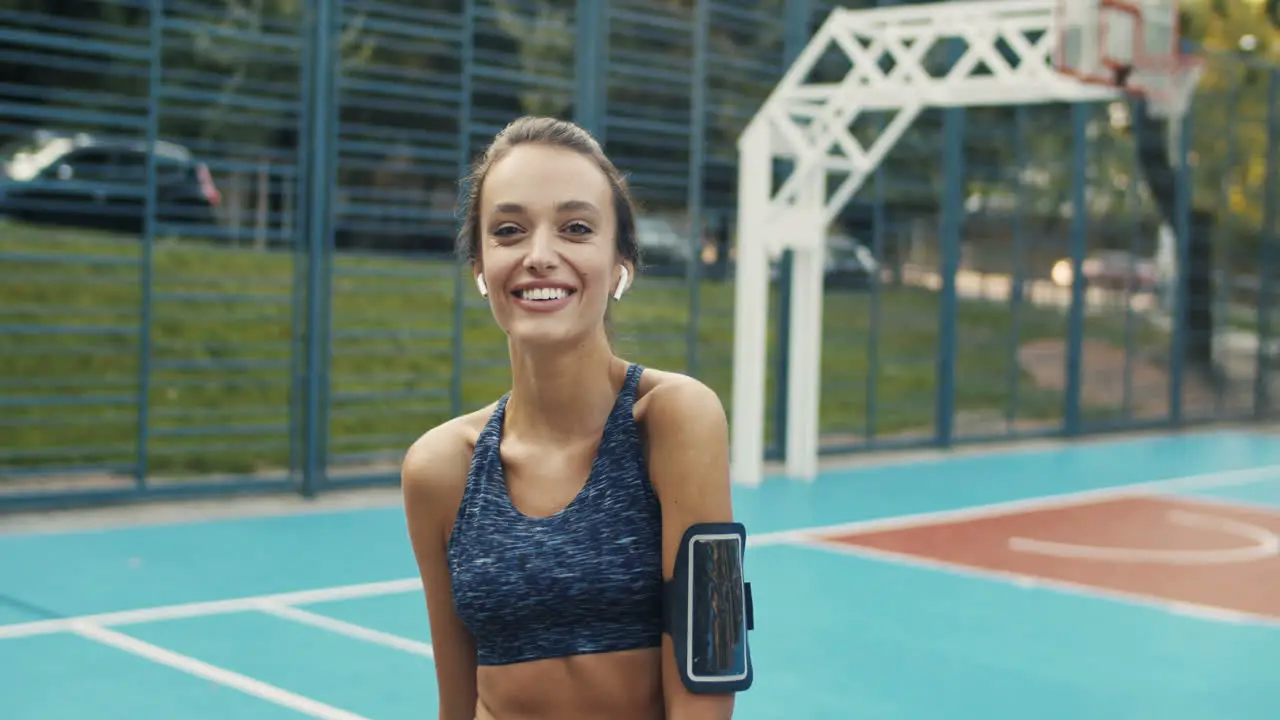 Portrait Of A Sporty Woman With Airpods Smiling Cheerfully At The Camera While Standing At Sport Court On A Summer Day