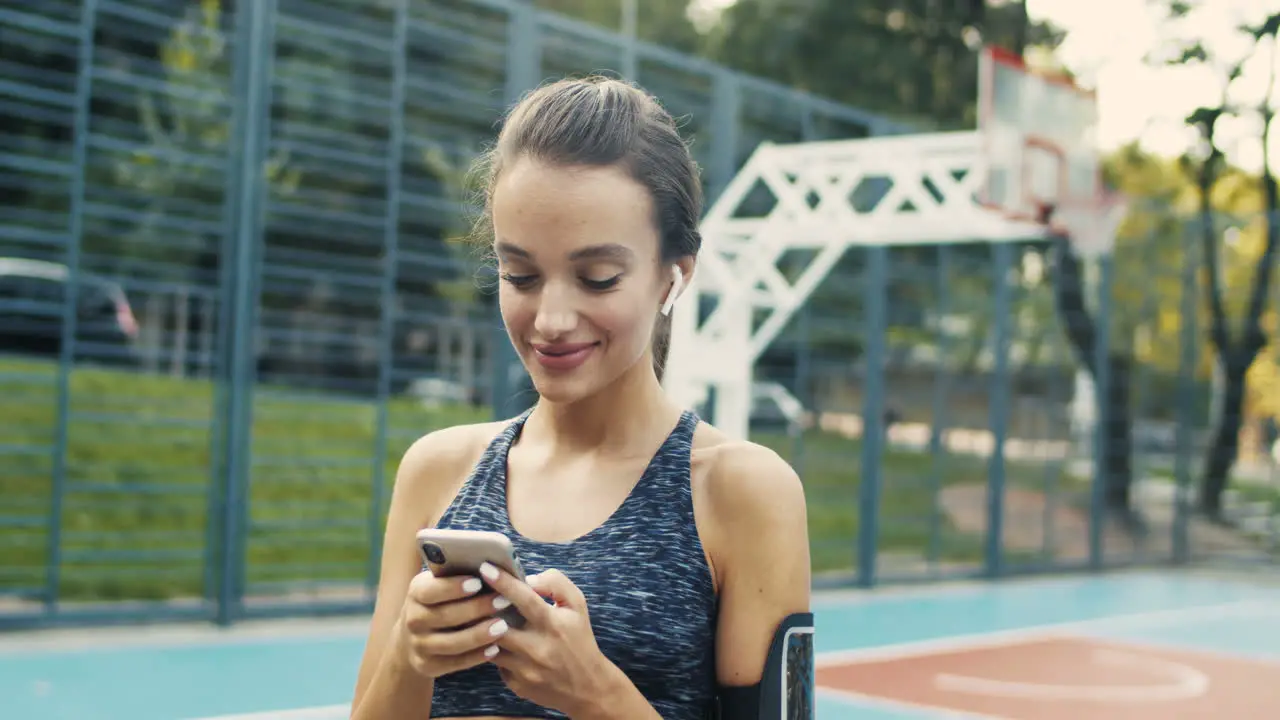 Sporty Girl With Airpods Laughing While Texting Message On Smartphone At Outdoor Court On A Summer Day