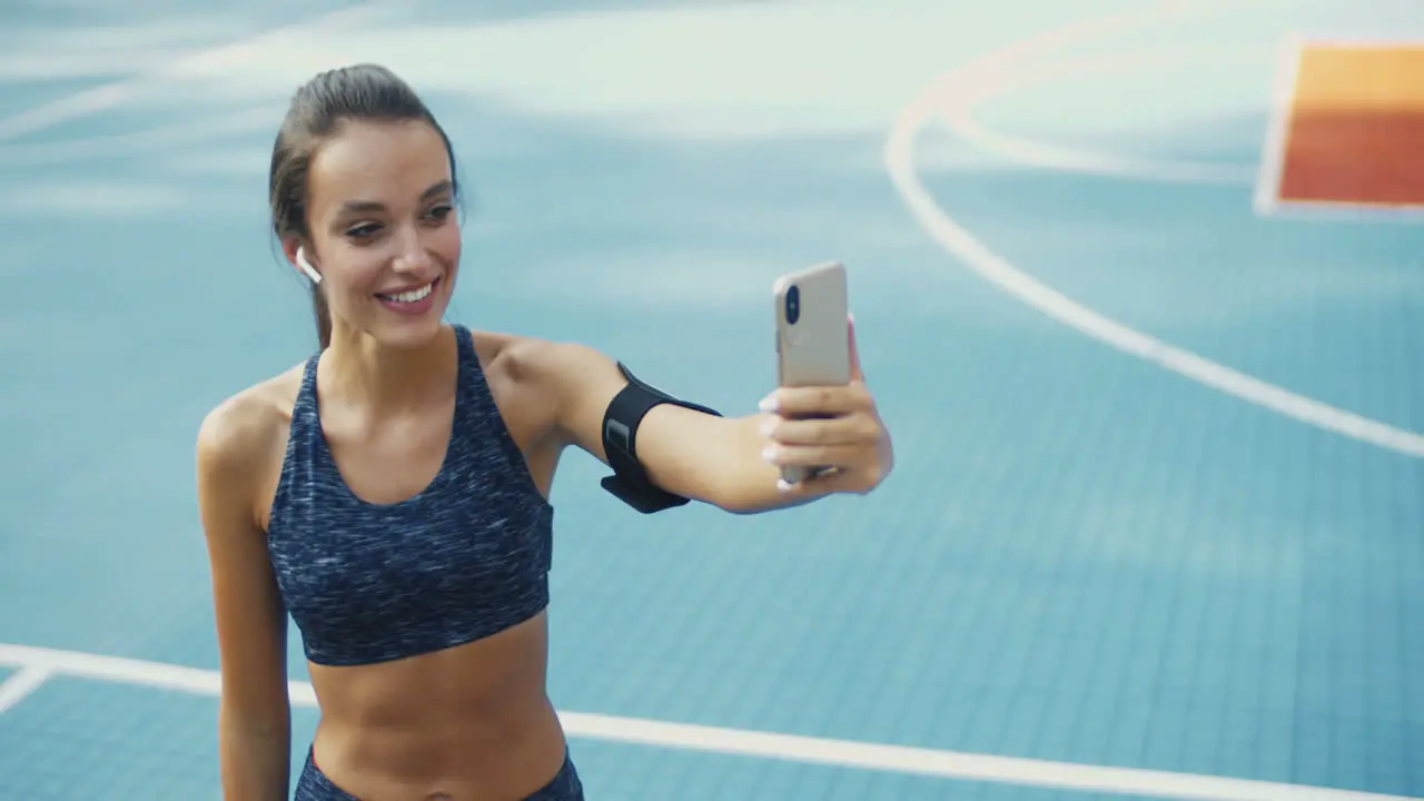 Cheerful Sporty Woman With Airpods Smiling And Showing Biceps To Smartphone Camera While Having Videochat At Sport Court