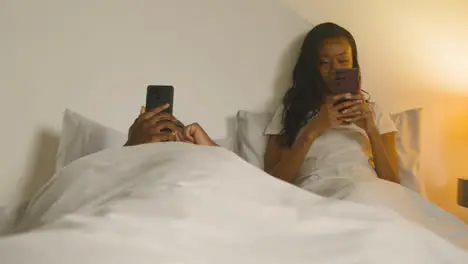 Couple With Relationship Problems At Home At Night Lying In Bed Both Looking At Mobile Phones