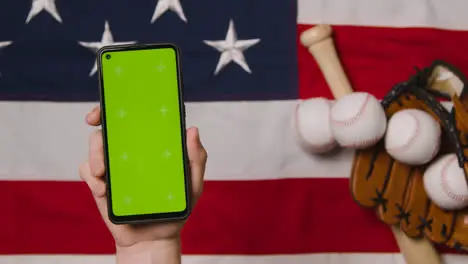 Person Holding Green Screen Mobile Phone Above Baseball Still Life With Bat Ball And Catchers Mitt On American Flag