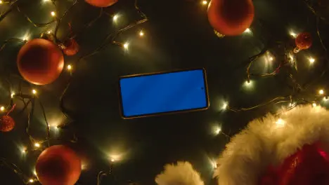 Overhead Shot Of Revolving Blue Screen Mobile Phone With Christmas Decorations Lights And Santa Hat 1