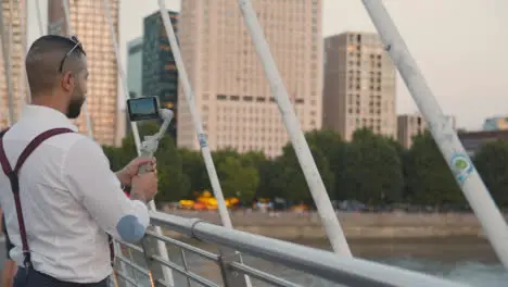 Man Recording Video On Mobile Phone Using Gimbal From Hungerford Bridge Over Thames London England UK