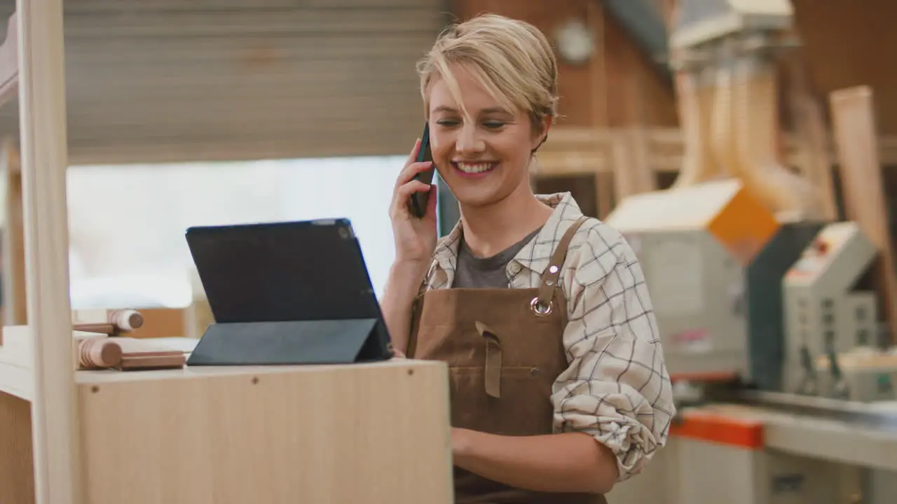 Female Apprentice Carpenter With Digital Tablet Working In Furniture Workshop Making Phone Call