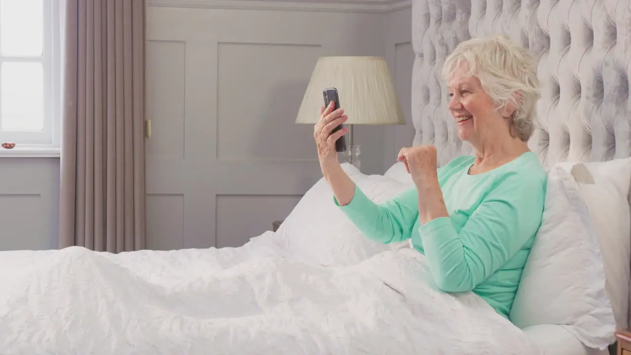 Senior Woman At Home Wearing Pyjamas In Bed Making Video Call On Mobile Phone