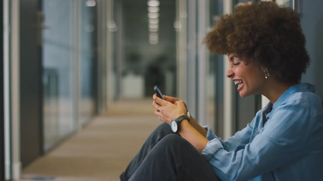 Young Businesswoman Sitting On Floor In Corridor Of Modern Office With Phone Celebrating Good News