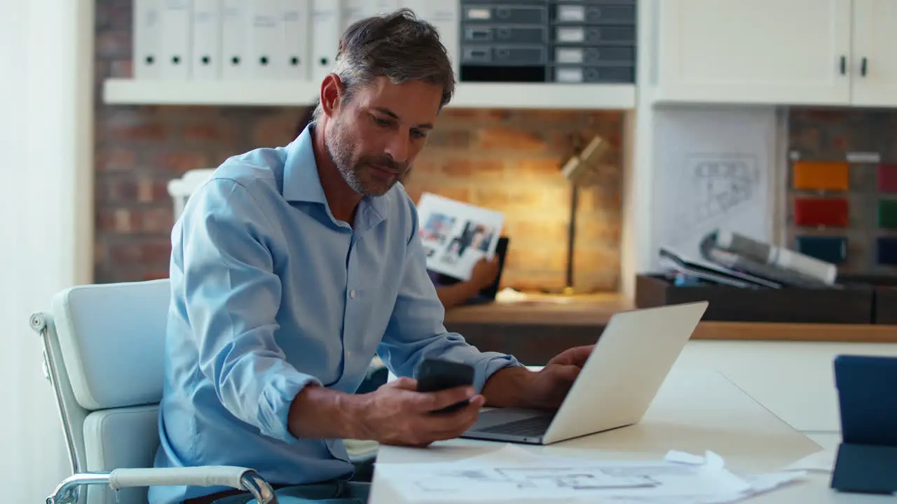 Mature Smiling Businessman Using Laptop Working At Desk In Office Taking Call On Mobile Phone