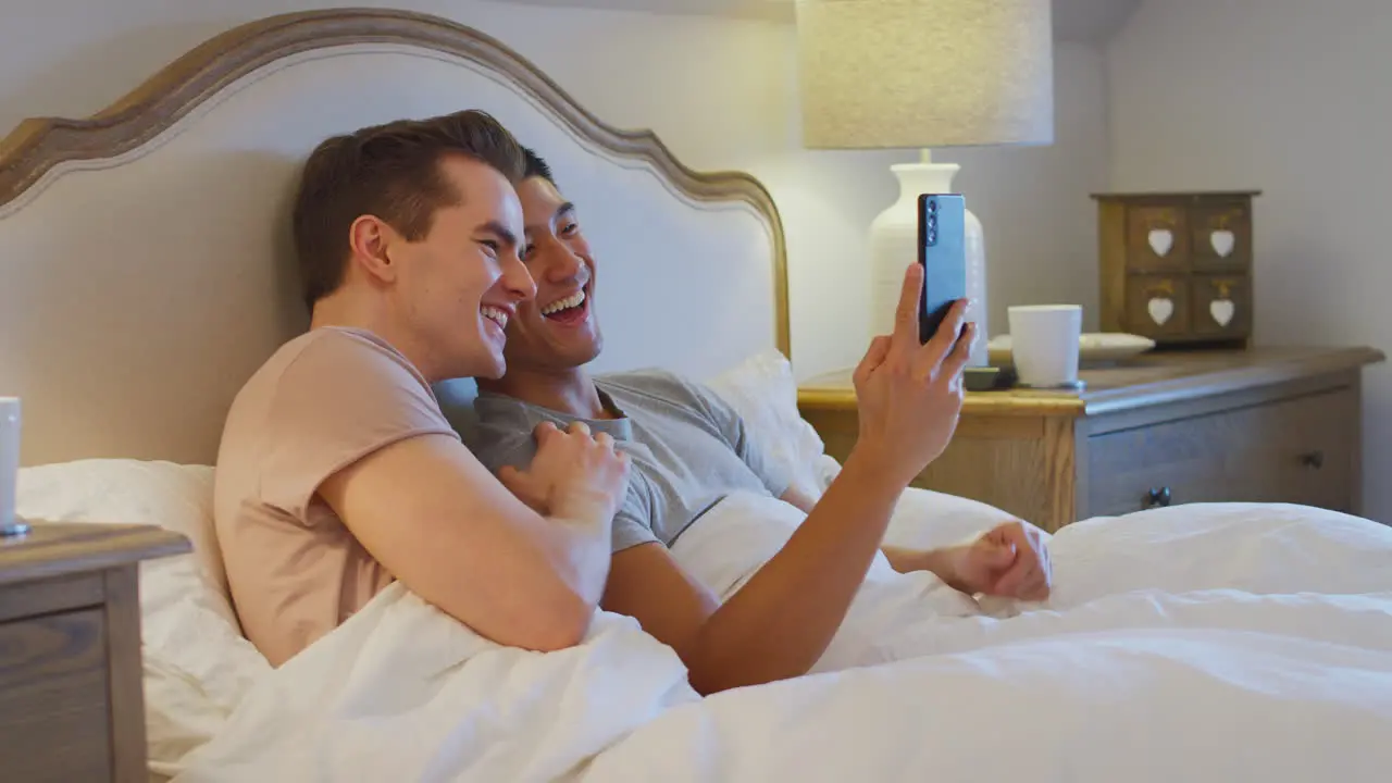 Same Sex Male Couple Lying In Bed At Home Making Video Call On Mobile Phone