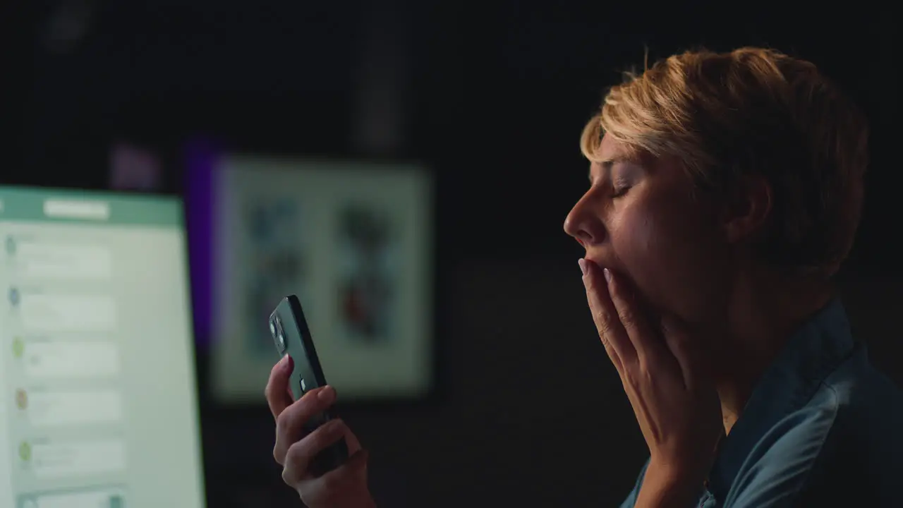 Face Of Tired Businesswoman Yawning Working Late In Office Using Mobile Phone Lit By Computer Screen