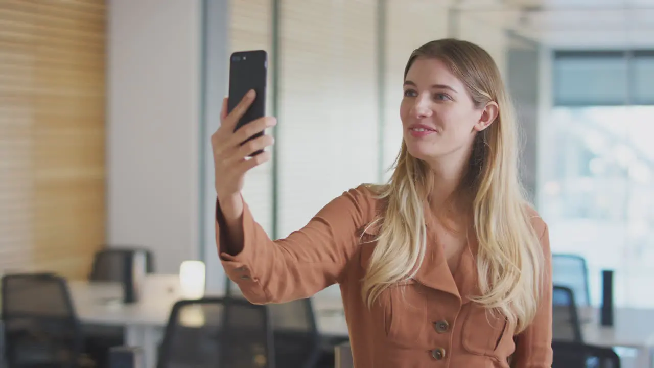 Businesswoman Having Video Chat On Mobile Phone Call Standing In Modern Open Plan Office