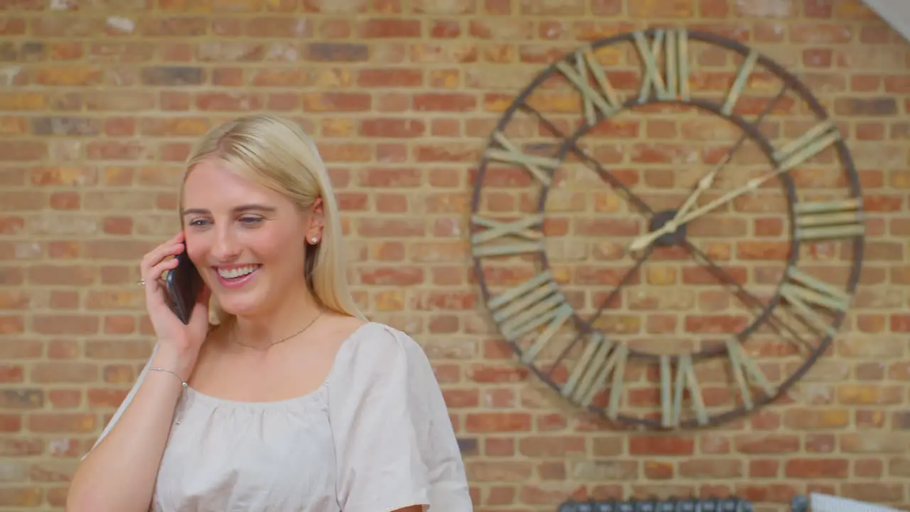 Young Woman Standing By Brick Wall With Clock At Home Talking With Friends On Mobile Phone