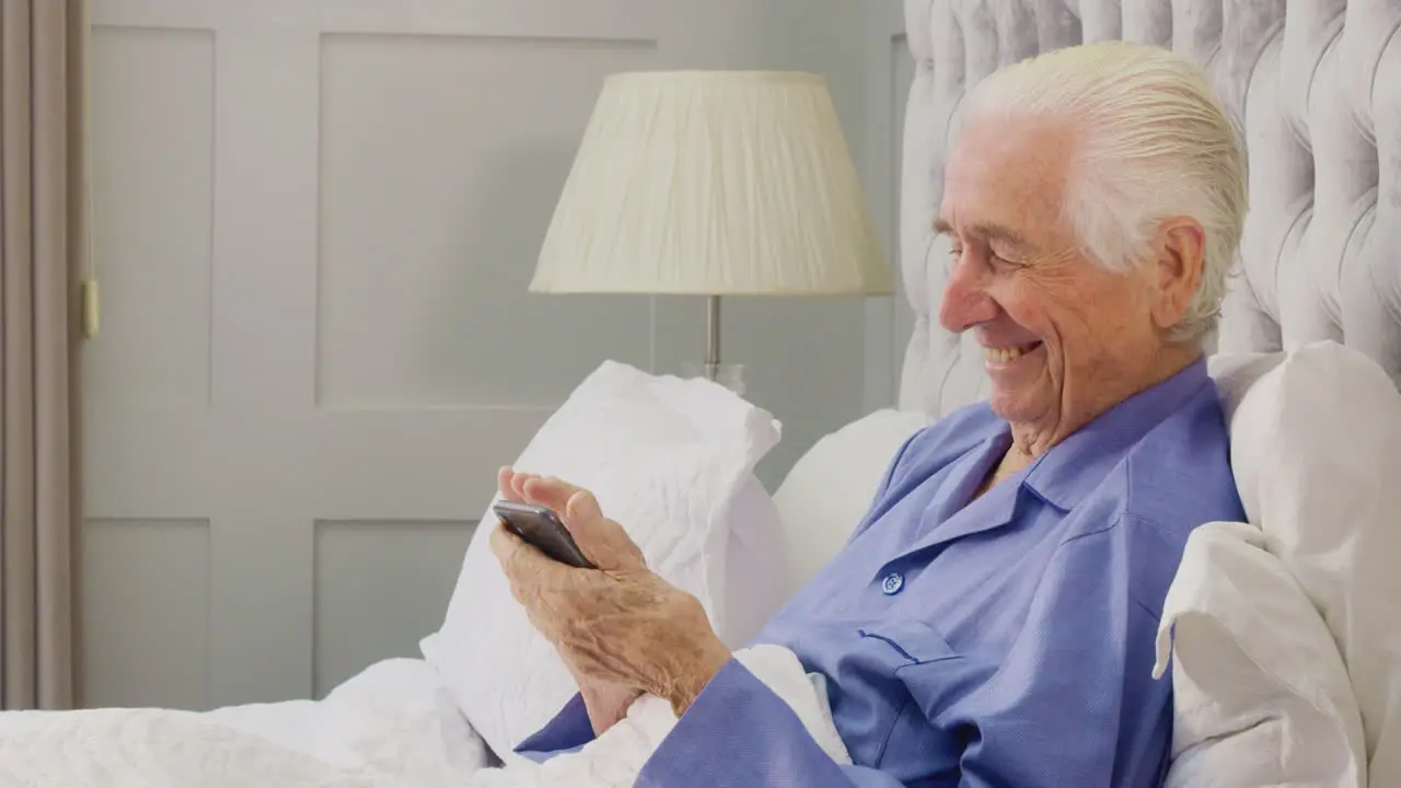 Smiling Senior Man At Home Wearing Pyjamas In Bed Using Mobile Phone