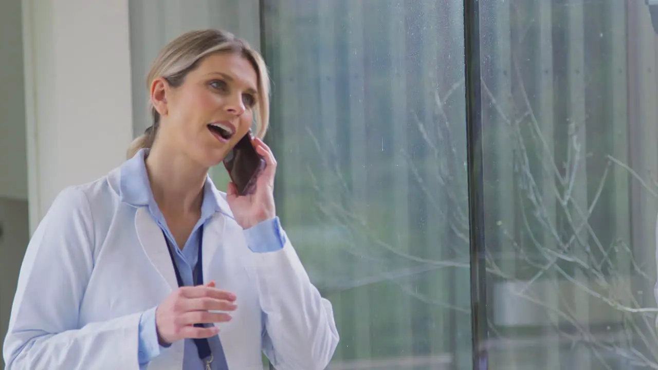 Female Doctor Wearing White Coat Standing In Hospital Corridor Talking On Mobile Phone