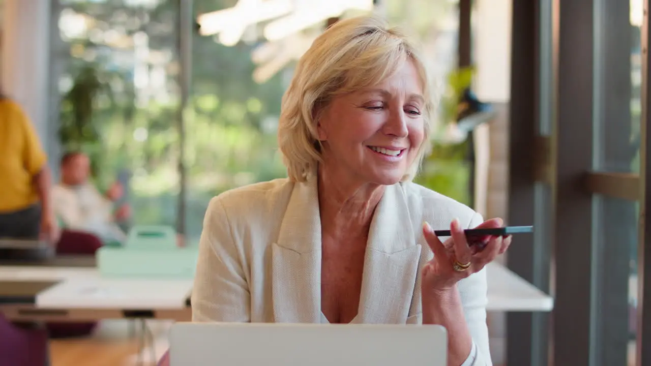 Mature Businesswoman Working On Laptop At Desk In Office Talking Into Mic Of Mobile Phone