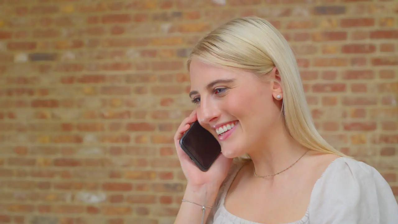 Young Woman Standing By Brick Wall At Home Talking With Friends On Mobile Phone